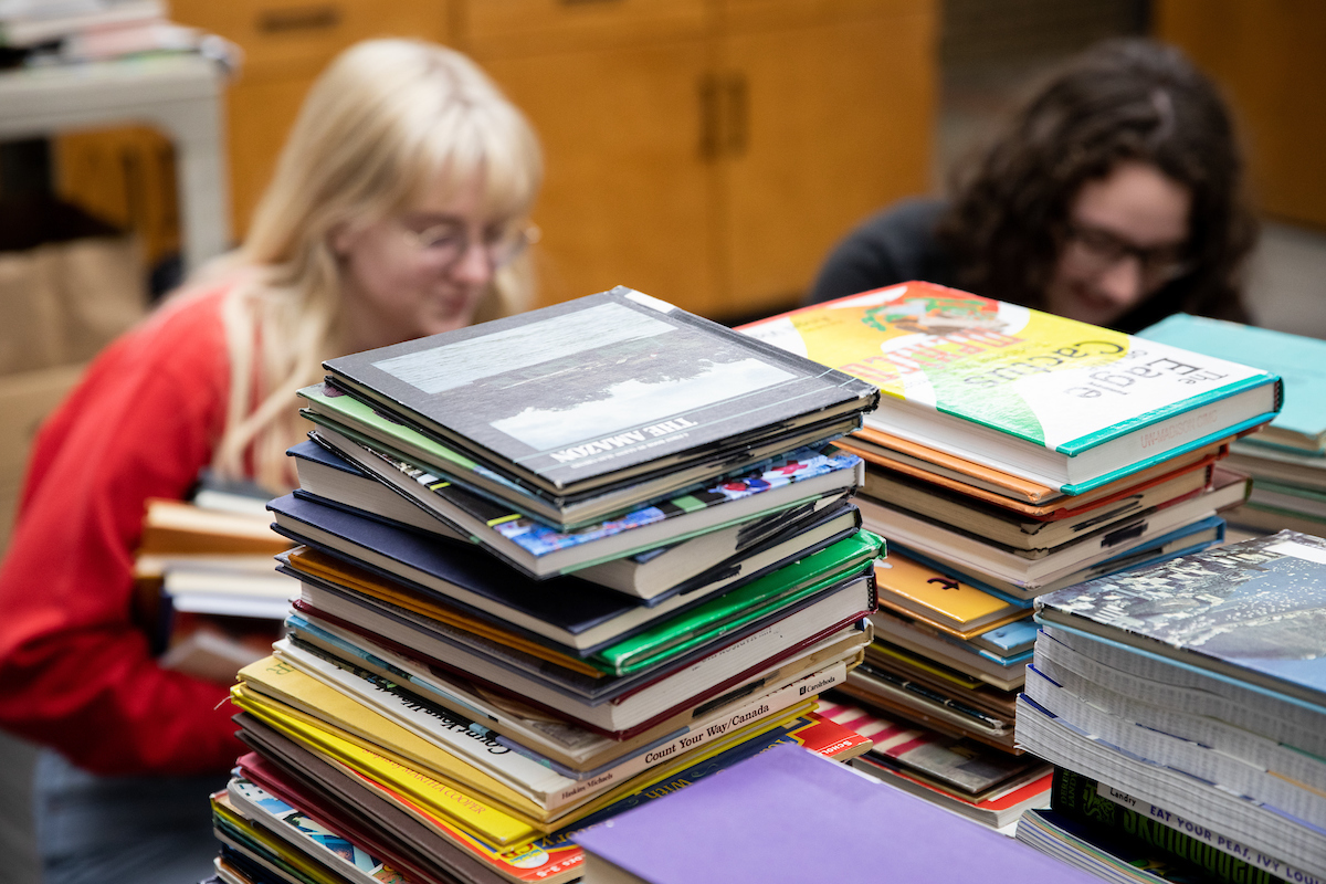 A stack of books in the foreground with two students in the background